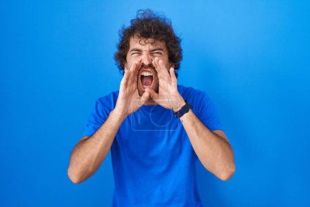 Téléchargez les photos : Jeune homme hispanique debout sur fond bleu criant en colère à haute voix avec les mains sur la bouche - en image libre de droit