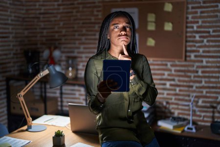 Photo for Young african american with braids working at the office at night thinking concentrated about doubt with finger on chin and looking up wondering - Royalty Free Image