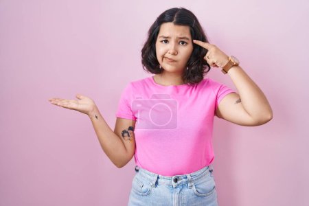 Photo for Young hispanic woman standing over pink background confused and annoyed with open palm showing copy space and pointing finger to forehead. think about it. - Royalty Free Image