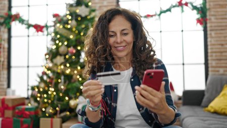 Foto de Mujer hispana de mediana edad comprando con smartphone y tarjeta de crédito celebrando la Navidad en casa - Imagen libre de derechos