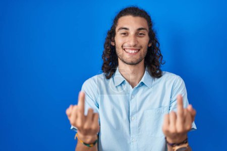 Foto de Young hispanic man standing over blue background showing middle finger doing fuck you bad expression, provocation and rude attitude. screaming excited - Imagen libre de derechos
