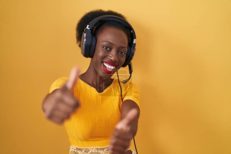 Foto de Mujer africana con el pelo rizado de pie sobre fondo amarillo con auriculares que aprueban hacer gesto positivo con la mano, pulgares hacia arriba sonriendo y feliz por el éxito. gesto ganador. - Imagen libre de derechos
