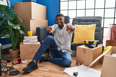 Foto de Hombre afroamericano sentado en el suelo en una casa nueva señalándote a ti y a la cámara con los dedos, sonriendo positivo y alegre - Imagen libre de derechos