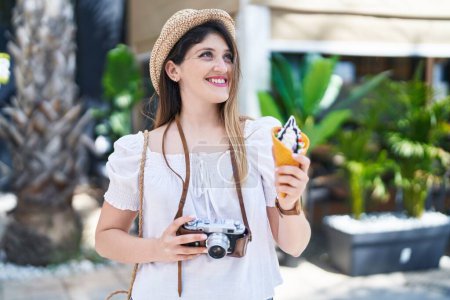 Foto de Young hispanic woman tourist holding camera eating ice cream at park - Imagen libre de derechos
