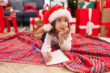 Photo for Adorable hispanic girl writing santa claus letter lying on floor by christmas tree at home - Royalty Free Image