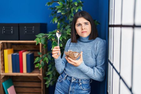 Foto de Mujer hispana joven comiendo cereales integrales saludables con una cuchara de expresión despistada y confusa. concepto de duda. - Imagen libre de derechos