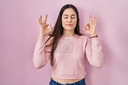 Photo for Young brunette woman standing over pink background relaxed and smiling with eyes closed doing meditation gesture with fingers. yoga concept. - Royalty Free Image