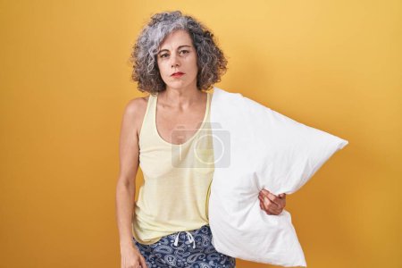 Téléchargez les photos : Femme d'âge moyen aux cheveux gris portant un pyjama étreignant l'oreiller somnolent et fatigué, épuisée par la fatigue et la gueule de bois, les yeux paresseux le matin. - en image libre de droit