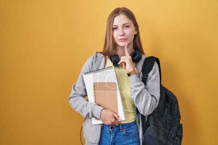 Foto de Joven mujer caucásica usando mochila de estudiante y sosteniendo libros pensando concentrado en la duda con el dedo en la barbilla y mirando hacia arriba preguntándose - Imagen libre de derechos