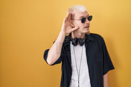 Téléchargez les photos : Jeune homme caucasien portant des lunettes de soleil debout sur fond jaune souriant avec la main sur l'oreille en écoutant une rumeur ou des commérages. concept de surdité. - en image libre de droit