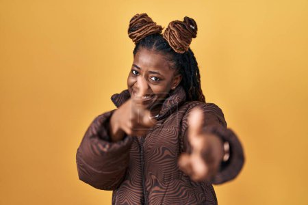 Foto de Mujer africana con el pelo trenzado de pie sobre el fondo amarillo señalando los dedos a la cámara con la cara feliz y divertida. buena energía y vibraciones. - Imagen libre de derechos