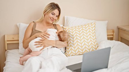 Foto de Madre e hija sentadas en la cama amamantando al bebé viendo películas en el portátil en el dormitorio - Imagen libre de derechos