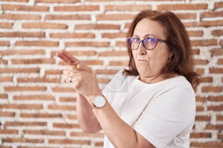 Foto de Mujer mayor con gafas de pie sobre la pared de ladrillos sosteniendo un arma simbólica con gesto de mano, jugando a matar armas de fuego, cara enojada - Imagen libre de derechos