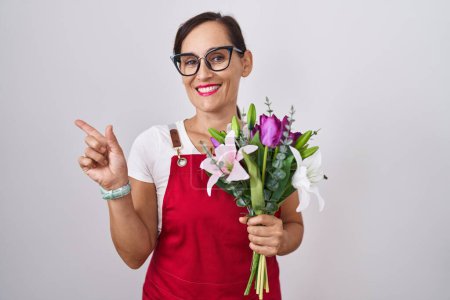 Foto de Mujer morena de mediana edad usando delantal trabajando en floristería sosteniendo ramo con una gran sonrisa en la cara, señalando con el dedo de la mano hacia el lado mirando a la cámara. - Imagen libre de derechos