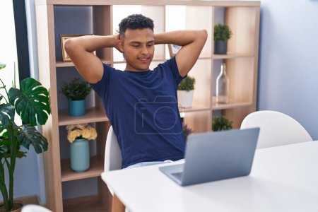 Téléchargez les photos : Jeune homme latin utilisant un ordinateur portable détendu avec les mains sur la tête à la maison - en image libre de droit