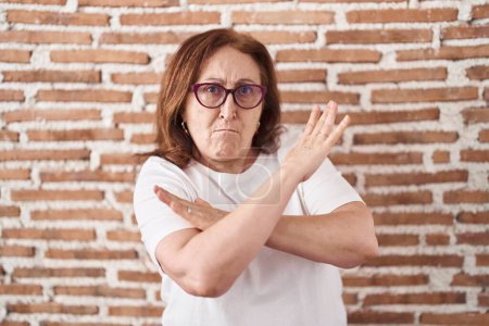 Foto de Mujer mayor con gafas de pie sobre ladrillos expresión rechazo de la pared cruzando brazos haciendo signo negativo, cara enojada - Imagen libre de derechos