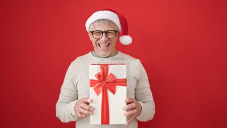 Foto de Hombre de pelo gris de mediana edad sosteniendo regalo con sombrero de Navidad sobre fondo rojo aislado - Imagen libre de derechos