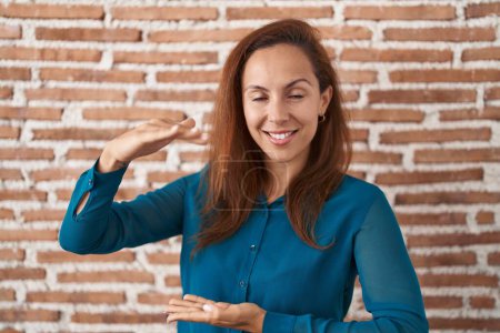 Téléchargez les photos : Femme brune debout sur le mur de briques geste avec les mains montrant grand et grand signe de taille, symbole de mesure. souriant en regardant la caméra. concept de mesure. - en image libre de droit