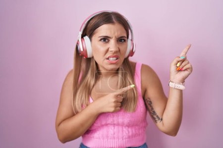 Foto de Mujer rubia joven escuchando música usando auriculares apuntando a un lado preocupada y nerviosa con ambas manos, preocupada y sorprendida expresión - Imagen libre de derechos
