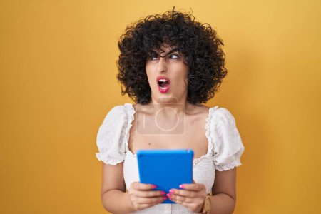 Photo for Young brunette woman with curly hair using touchpad over yellow background angry and mad screaming frustrated and furious, shouting with anger. rage and aggressive concept. - Royalty Free Image