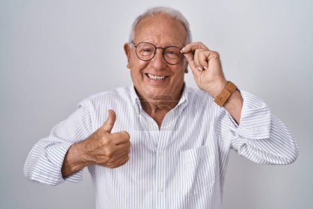 Foto de Hombre mayor con el pelo gris sosteniendo gafas con la mano sonriendo feliz y positivo, pulgar hacia arriba haciendo excelente y signo de aprobación - Imagen libre de derechos