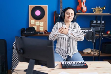 Photo for Young modern girl with blue hair at music studio wearing headphones smiling with hands on chest, eyes closed with grateful gesture on face. health concept. - Royalty Free Image