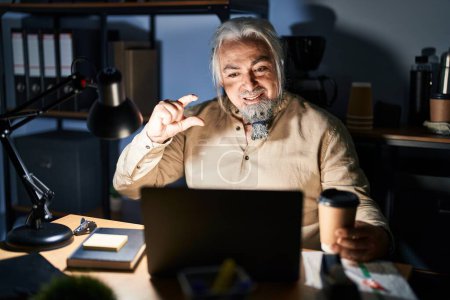 Photo for Middle age man with grey hair working at the office at night smiling and confident gesturing with hand doing small size sign with fingers looking and the camera. measure concept. - Royalty Free Image