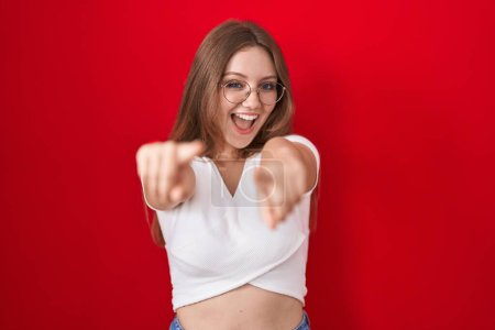 Téléchargez les photos : Jeune femme caucasienne debout sur fond rouge pointant vers vous et la caméra avec les doigts, souriant positif et joyeux - en image libre de droit