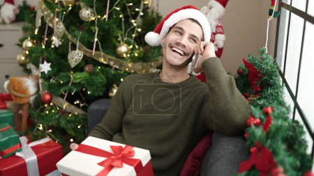 Photo for Young hispanic man talking on smartphone sitting on sofa holding christmas gift at home - Royalty Free Image