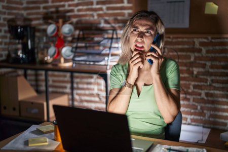 Photo for Young beautiful woman working at the office at night speaking on the phone thinking worried about a question, concerned and nervous with hand on chin - Royalty Free Image