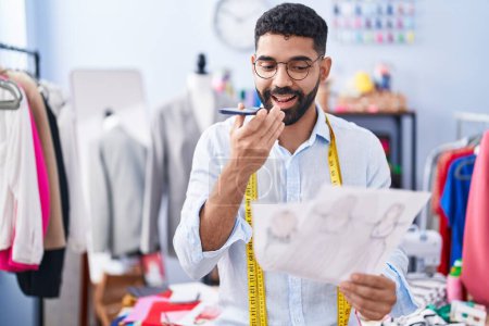 Téléchargez les photos : Young arab man tailor talking on smartphone looking clothing design at tailor shop - en image libre de droit