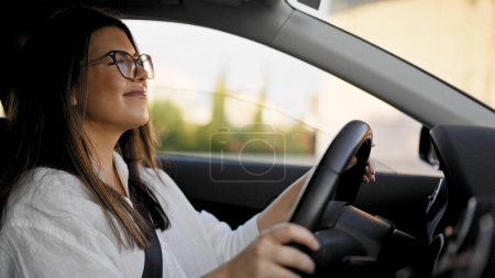 Foto de Joven mujer hispana hermosa conduciendo un coche sonriendo con gafas en la carretera - Imagen libre de derechos