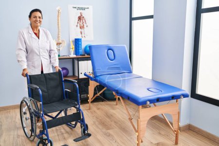 Téléchargez les photos : Femme hispanique d'âge moyen avec fauteuil roulant à la clinique de physiothérapie regardant positif et heureux debout et souriant avec un sourire confiant montrant des dents - en image libre de droit