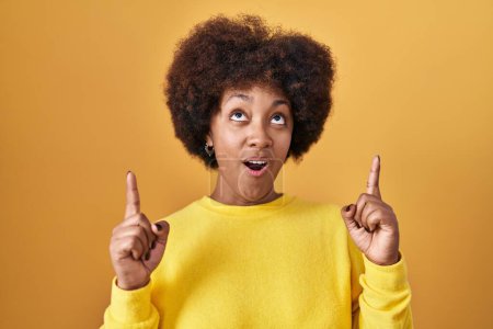 Foto de Joven mujer afroamericana de pie sobre fondo amarillo asombrada y sorprendida mirando hacia arriba y señalando con los dedos y los brazos levantados. - Imagen libre de derechos