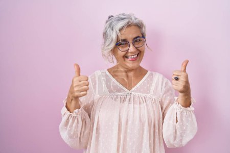 Téléchargez les photos : Femme d'âge moyen avec les cheveux gris debout sur fond rose signe de succès faisant geste positif avec la main, pouces levés souriant et heureux. expression joyeuse et geste gagnant. - en image libre de droit