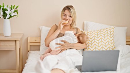 Foto de Madre e hija amamantando bebé viendo la película en el ordenador portátil comiendo croissant en el dormitorio - Imagen libre de derechos