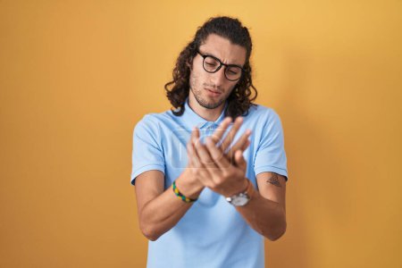 Téléchargez les photos : Jeune homme hispanique debout sur fond jaune souffrant de douleur aux mains et aux doigts, inflammation de l'arthrite - en image libre de droit