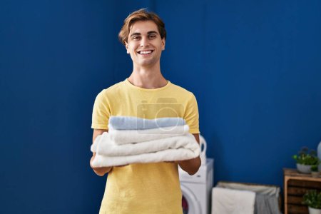 Téléchargez les photos : Young caucasian man smiling confident holding folded towels at laundry room - en image libre de droit