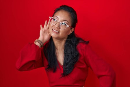 Foto de Mujer joven asiática de pie sobre fondo rojo sonriendo con la mano sobre el oído escuchando un rumor o chismes. concepto de sordera. - Imagen libre de derechos