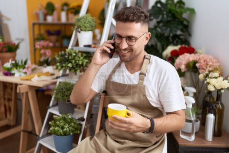 Téléchargez les photos : Jeune homme hispanique fleuriste boire une tasse de café parler sur smartphone à la boutique de fleurs - en image libre de droit