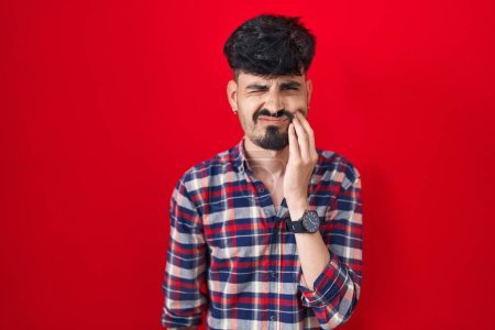 Téléchargez les photos : Jeune homme hispanique avec la barbe debout sur fond rouge touchant la bouche avec la main avec une expression douloureuse à cause de maux de dents ou de maladies dentaires sur les dents. dentiste - en image libre de droit