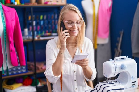 Téléchargez les photos : Jeune femme blonde tailleur parlant sur smartphone lecture carnet au studio de couture - en image libre de droit
