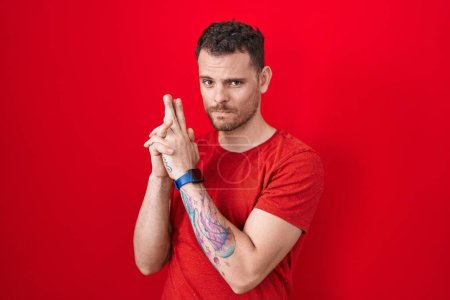 Photo for Young hispanic man standing over red background holding symbolic gun with hand gesture, playing killing shooting weapons, angry face - Royalty Free Image