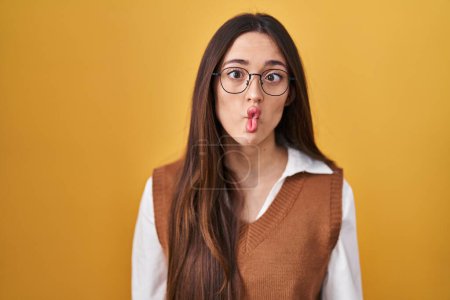 Téléchargez les photos : Jeune femme brune debout sur fond jaune portant des lunettes faisant visage de poisson avec des lèvres, geste fou et comique. expression drôle. - en image libre de droit