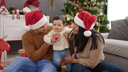 Téléchargez les photos : Couple et fils tenant boule de décoration assis sur le sol par arbre de Noël à la maison - en image libre de droit