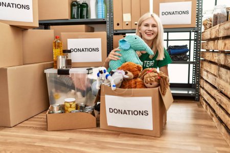 Téléchargez les photos : Jeune femme blonde volontaire souriant confiant emballage jouets sur carton boîte au centre de charité - en image libre de droit