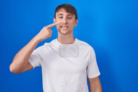 Téléchargez les photos : Homme blond caucasien debout sur fond bleu pointant avec le doigt de la main sur le visage et le nez, souriant gai. concept de beauté - en image libre de droit