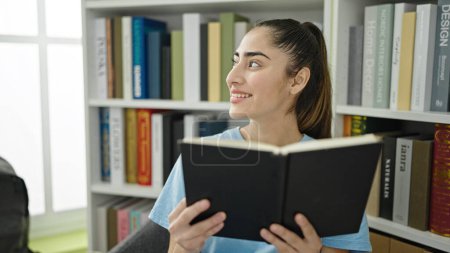 Téléchargez les photos : Jeune belle hispanique étudiant livre de lecture assis sur la chaise à l'université de bibliothèque - en image libre de droit