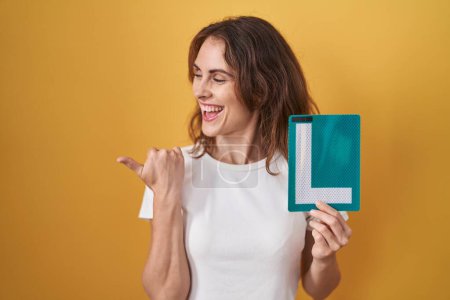 Photo for Beautiful brunette woman holding l sign for new driver pointing thumb up to the side smiling happy with open mouth - Royalty Free Image