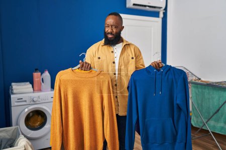 Foto de Hombre afroamericano sosteniendo ropa limpia en perchas en la lavandería expresión despistada y confusa. concepto de duda. - Imagen libre de derechos